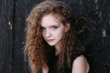 Siyah elbiseli kızıl saçlı kız koyu dokulu bir duvarın yanında.