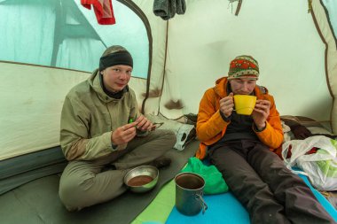 Dağlarda yürüyüş yaparken çadırda yemek yiyen iki adam.