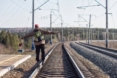 Sırt çantalı genç bir adam güneşli bir günde tren raylarında yürüyor.