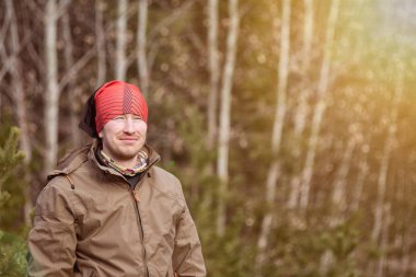Kırmızı şapkalı ve sıcak elbiseli bir adam güneşli bir günde ormanda yürüyor. 