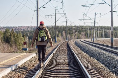 Sırt çantalı genç bir adam güneşli bir günde tren raylarında yürüyor.
