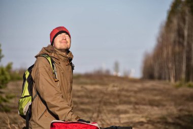 Kırmızı şapkalı ve sıcak elbiseli bir adam güneşli bir günde ormanda yürüyor. 
