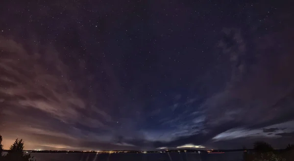 Astrofotoğrafçılık. Gece şehre yakın bir nehirdir, karşı kıyıda şehrin ışıklarını görebilirsiniz. Gökyüzünde, bulutlar renkli ve birçok yıldız var..