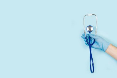 Açık mavi arkaplanda steteskop tutan eldiven ve fotokopi alanı olan bir el.