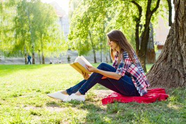 Parkta ağaç yakınındaki çimenlerin üzerinde günbatımında oturup kitap okuma gözlüğü olan güzel kadın.