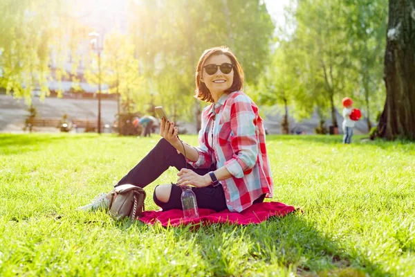 在耳机上听音乐的成熟女人 坐在草地上的公园 休息享受自然 — 图库照片