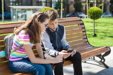 Erkek ve kız çocuk oyun, okumak, smartphone vasıl bak. Bankta kentsel arka plan.