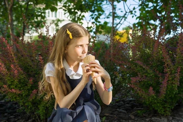 Schoolgirl 8 years old eating ice cream. Blond girl in school uniform, background of green trees, yard.