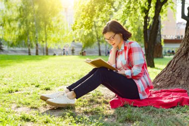 Parkta ağaç yakınındaki çimenlerin üzerinde günbatımında oturup kitap okuma gözlüğü olan güzel olgun kadın.