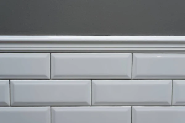 Gray painted wall, part of the wall is covered tiles small white glossy brick, ceramic decorative molding tiles. Fragment of the walls of the bathroom, toilet, kitchen. Interior details close-up.