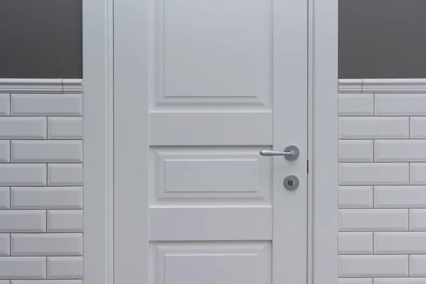 White door in the bathroom toilet room. Background gray painted wall covered with decorative ceramic tiles with white glossy bricks. Interior details close-up