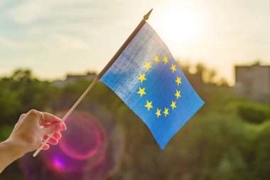 Hand holds European Union flag in an open window. Background blue sky, silhouette of the city, sunset. clipart