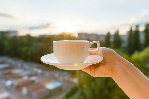 女性手捧着白杯和茶托 背景黄昏日落 城市剪影 — 图库照片