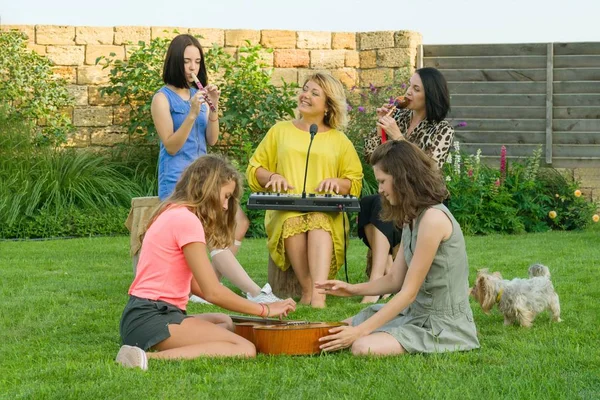 A családi szórakozás, a két anya, tizenéves lánya énekel és hangszerek, használ családi zenekar, ült a gyepen a ház közelében. — Stock Fotó