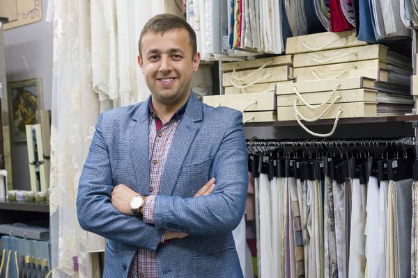 Portrait of happy businessman owner with crossed arms in interior fabrics store, background fabric samples. Small business home textile shop