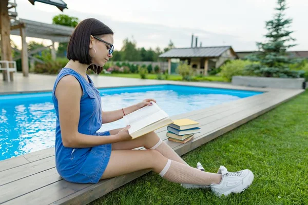 戴眼镜的十几岁的女孩读了一本书 背景游泳池 房子附近的草坪 青少年 — 图库照片