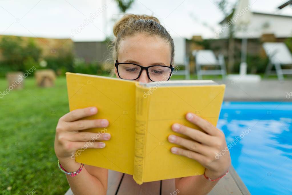 Young girl in glasses near the pool with a pile of books, reading book. Education, summer, knowledge