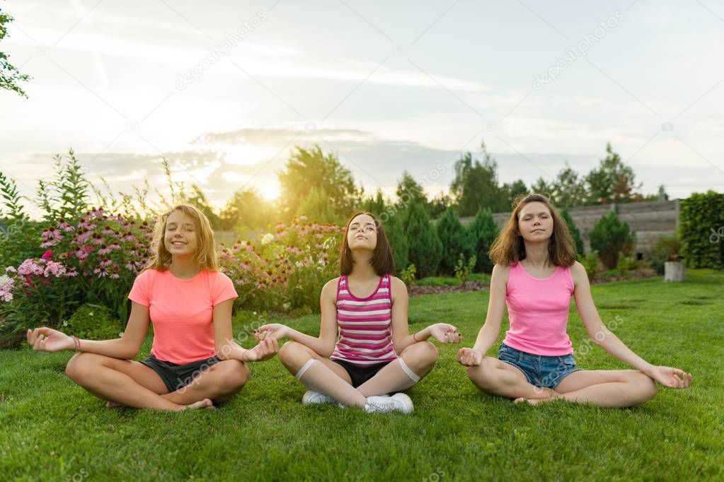 Group of young teenage girls practice yoga, meditate, sit in a lotus position on the grass near the house.