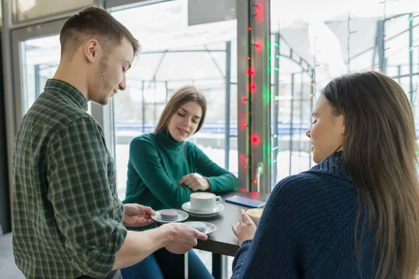 Café, saison d'hiver, deux jeunes femmes assises à la table avec une tasse de café, et barista mâle avec des macarons sur l'assiette souriant et parlant — Photo