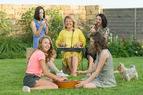 Rodzina jest dobra zabawa, dwie matki z nastoletnich córek są śpiewa i za pomocą instrumentów muzycznych, zespół muzyczny dla rodzin z dziećmi, siedząc na trawnik w pobliżu domu — Zdjęcie stockowe