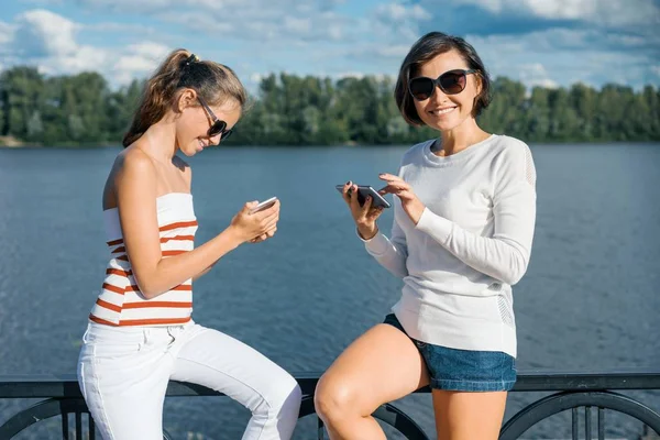 Moeder en dochter lopen in een zomer-park in de buurt van de rivier, met behulp van mobiele telefoons, praten en lachen — Stockfoto