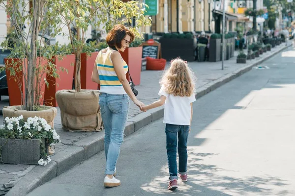 Мама с дочкой идут по улице, держась за руки, и вид сзади. Солнечный летний день в городе — стоковое фото