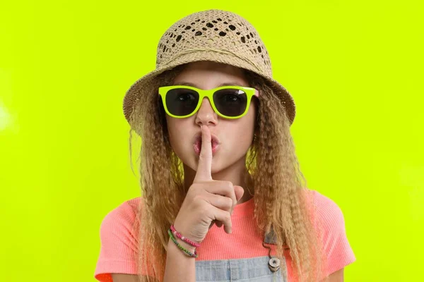 Porträt eines Teenie-Mädchens mit Sonnenbrille und Strohhut mit dem Zeigefinger in der Nähe der Lippen, zeigt Schweigezeichen, geheime Geste, gelber Studiohintergrund — Stockfoto