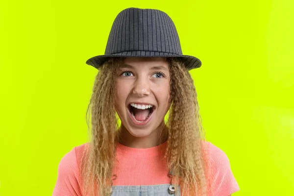 Sorprendida chica adolescente feliz con el pelo rizado en el sombrero mirando a la cámara con la boca abierta sobre fondo de estudio amarillo — Foto de Stock