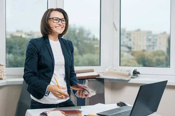 Innenarchitektin am Arbeitsplatz mit Stoffmustern, Laptop. Lächelnde Frau im Sakko, Brille zur Seite gerichtet, während sie neben dem Schreibtisch steht — Stockfoto