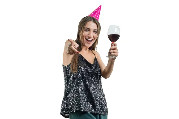 Jeune belle femme brune souriante avec un verre de vin rouge et portant un chapeau de fête pointe son doigt vers vous, isolée sur fond blanc. Fête, boissons, vacances et concept de célébration . — Photo