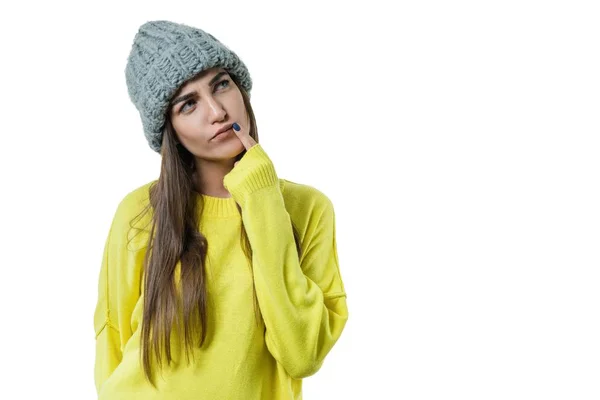 Mooie jongedame in de gele trui en grijze grote lus gebreide beanie muts, doordachte houden een vinger in de buurt van haar lippen, op witte geïsoleerde achtergrond — Stockfoto