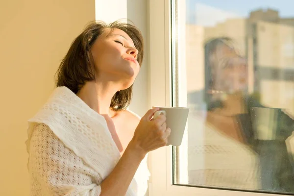 Mujer madura de pie cerca de la ventana con una taza de bebida caliente bajo una manta de lana de punto caliente se ve soñadoramente por la ventana, estilo otoño invierno — Foto de Stock