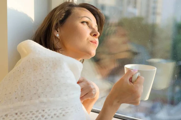 Mature woman standing near the window with cup of hot drink under warm knitted woolen blanket looks dreamily out the window, autumn winter style