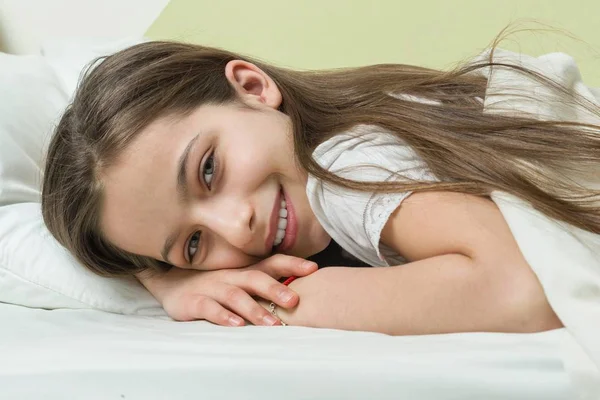 Крупным планом лицо красивой улыбающейся маленькой девочки, лежащей в постели на подушке — стоковое фото