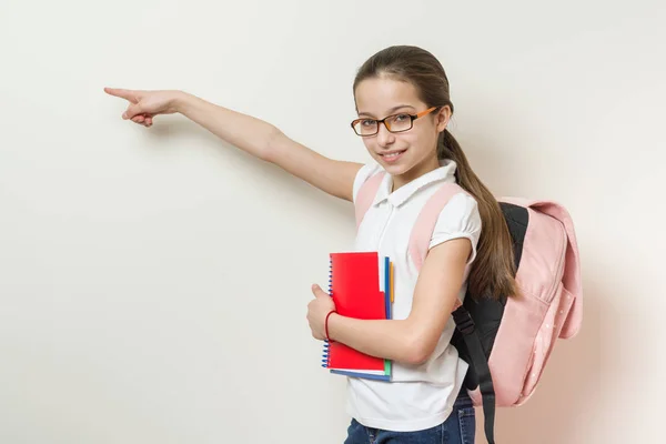 Μαθήτρια κορίτσι 10 ετών με ένα σακίδιο και βιβλία σημεία ένα δάχτυλο σε ένα κενό τοίχο, Χώρος για κείμενο — Φωτογραφία Αρχείου