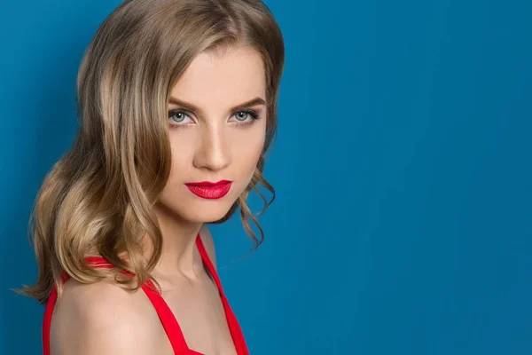 Retrato de belleza de una joven rubia con labios rojos brillantes, ojos azules, vestido rojo sobre fondo azul, espacio para copiar — Foto de Stock