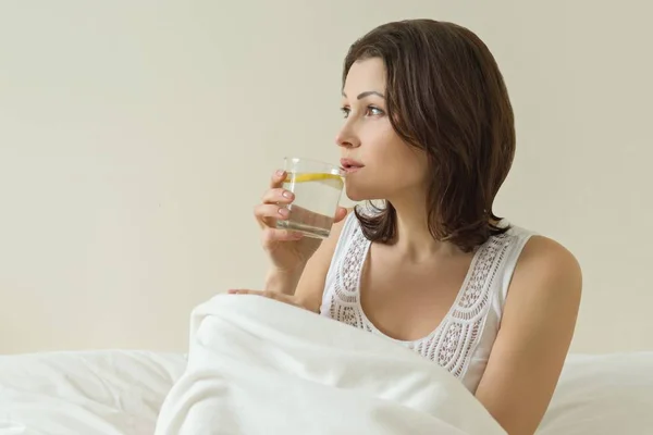 Middelbare leeftijd vrouw geniet van water met een citroen zitten in bed. Eerste ochtend drinken, antioxidant drink — Stockfoto