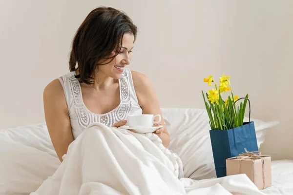 Mellersta år kvinna är nöjd med gåva, blombukett, sitter i morgon i sängen med kopp kaffe. Känslor av lycka, glädje, överraskning — Stockfoto