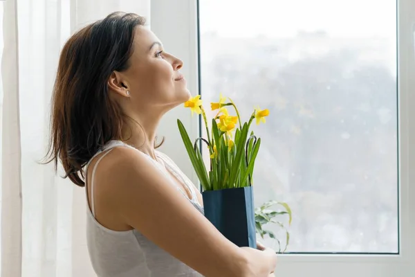 Glada leende mogen kvinna med bukett av gula blommor stående hemma nära fönstret vårsäsongen — Stockfoto