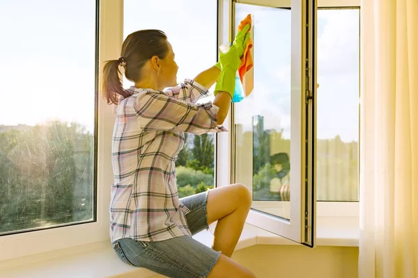 Frau mit Gummischutzhandschuhen, Lappen und Sprühwaschmittel blickt in ein sauber gewaschenes Fenster — Stockfoto
