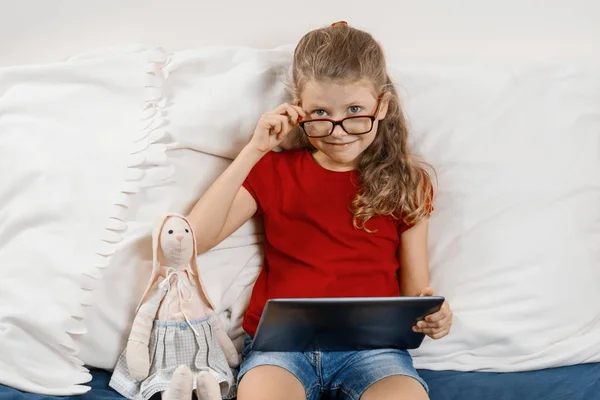 Маленькая симпатичная девочка в очках сидит дома в постели с игрушкой и цифровой планшет, отдых и образование дома. Улыбающийся ребенок смотрит в камеру без очков — стоковое фото