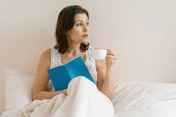 Mature woman at home resting in bed with a cup of fresh morning coffee and reading a book. Female looks in profile Stock Picture