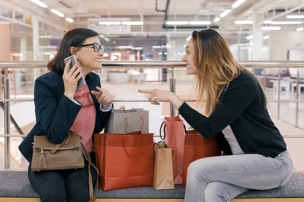 Две счастливые взрослые женщины подруги с сумками для покупок, женщины разговаривают сидя в торговом центре, со многими покупками — стоковое фото