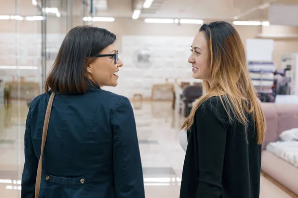 Две взрослые красивые улыбающиеся женщины в профиле, разговаривающие женщины крупным планом, задний план в помещении — стоковое фото