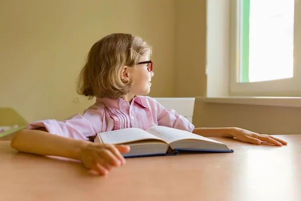 Κουρασμένος μικρό κορίτσι φοιτητής κοιτάζει έξω από το παράθυρο ενώ κάθεται στο γραφείο της με ένα μεγάλο βιβλίο. Σχολείο, παιδεία, γνώση και παιδιά — Φωτογραφία Αρχείου