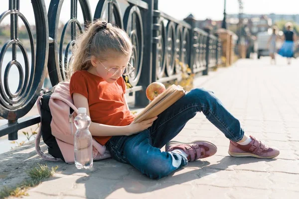 На відкритому повітрі портрет маленької школярки з книгою, дівчинки-дитини 7, 8 років з окулярами рюкзак читання підручник — стокове фото