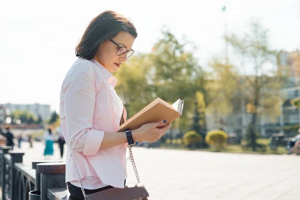 Outdoor portrait of mature caucasian woman 40, 45 years old with glasses reading book. — Stock Photo, Image