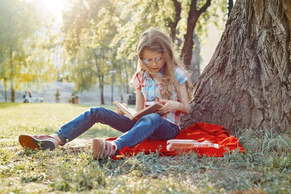 Девочка в очках читает книгу в парке, на траве возле дерева — стоковое фото