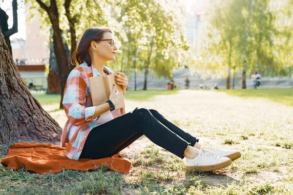 Hermosa mujer madura con gafas libro de lectura sentado en la hierba cerca del árbol en el parque al atardecer . —  Fotos de Stock