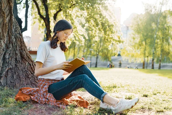 Uczeń dziewczyna w okularach, czytanie książki w parku, siedząc pod drzewem na trawie — Zdjęcie stockowe